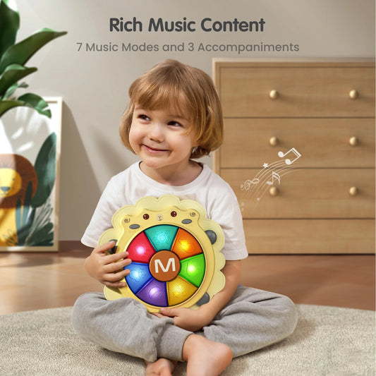 Smiling child sitting on the floor holding a sheep-shaped baby musical drum toy with colorful light-up buttons. The toy features 7 music modes and 3 accompaniments, providing rich music content to engage children in interactive play and early music education.