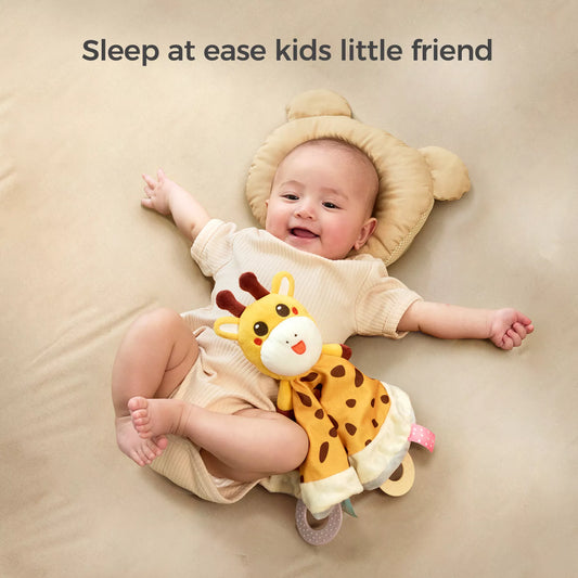 A smiling baby lying on a bed with a bear-shaped pillow, holding a cute giraffe baby security blanket. The text above says, "Sleep at ease kids little friend."