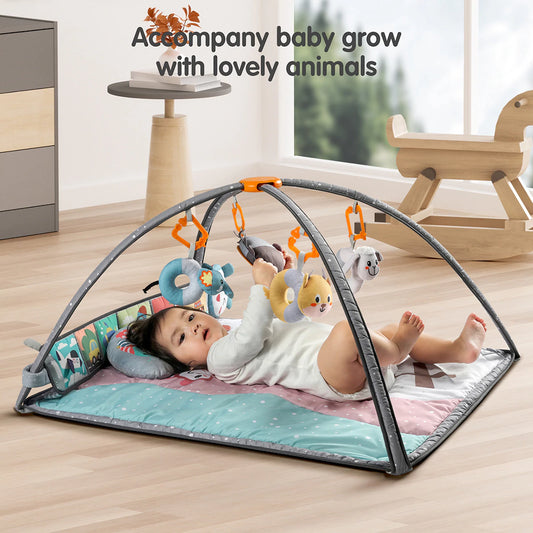 Baby lying on a colorful padded play mat with grey arches, reaching for hanging plush rattles including an elephant, lion, cat, and lamb. Side cloth book panel with animal illustrations for added interaction.
