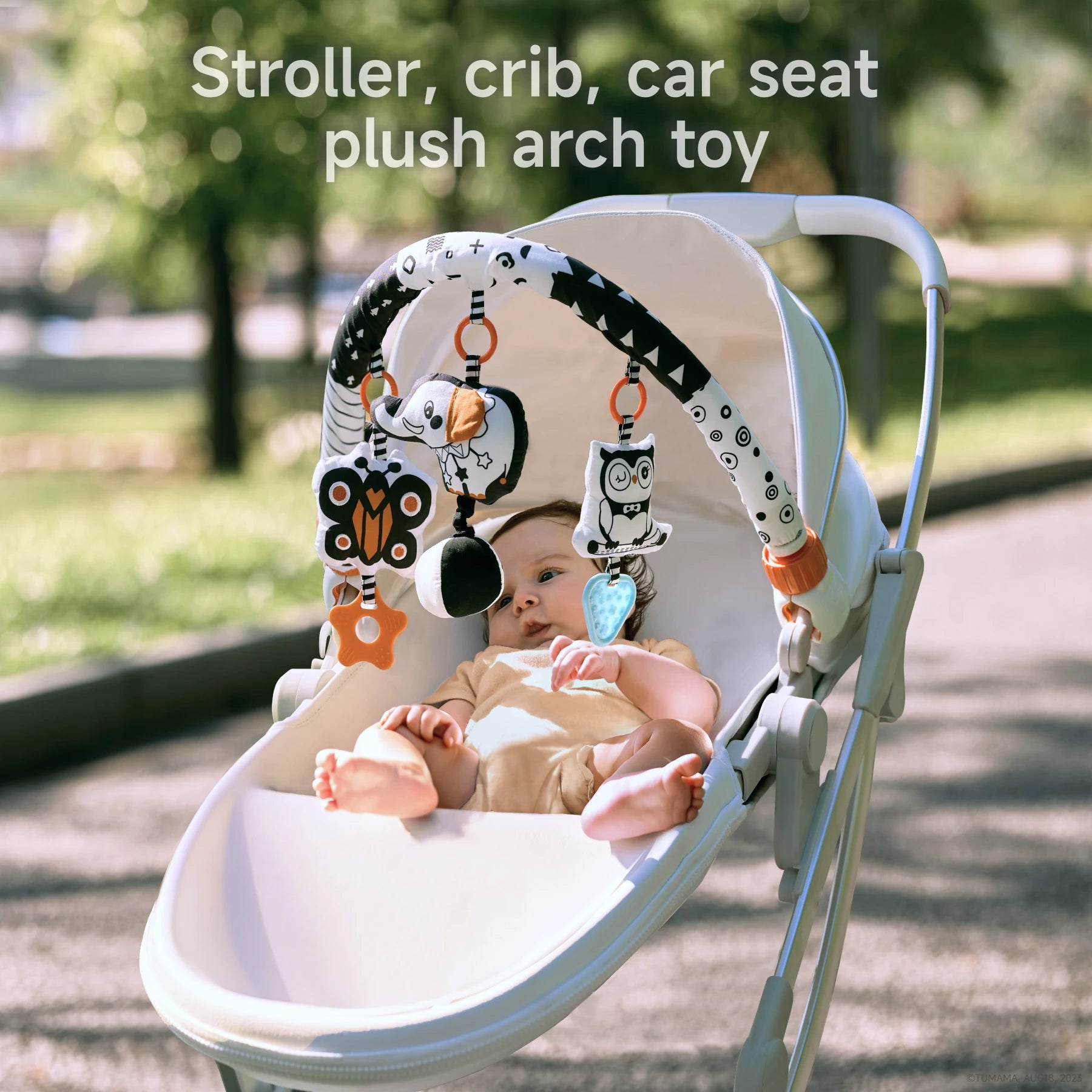 Baby lying in a white stroller outdoors with a black and white plush arch toy attached overhead. The toy features colorful hanging plush characters including a dog, owl, and butterfly, designed for use with strollers, cribs, and car seats.