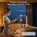 In a dim nursery a mother stands beside a white crib holding her smartphone to connect via Bluetooth (‘T.K. Crib Mobile Connected’) to a clamp-on musical mobile overhead, which features black-and-white plush woodland animals and projects light above her smiling baby.