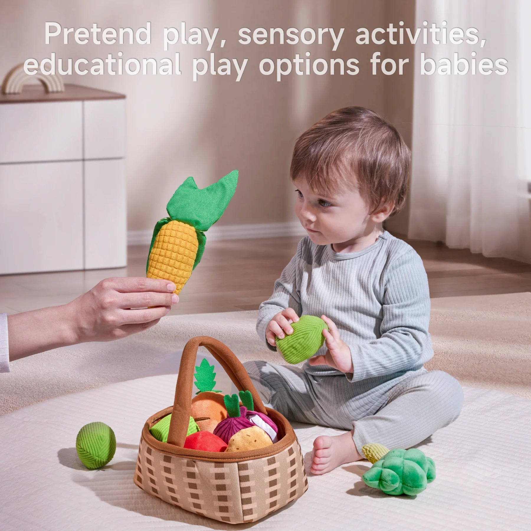 A hand holds a felted corn cob toy above a seated child’s head, who is grasping a green felted piece. Nearby, a basket overflows with assorted colorful felted vegetables on a light carpet.