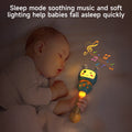 Sleeping baby holding a blue bunny-shaped musical rattle with teether ears and a glowing handle. The toy emits soft light and colorful musical notes, creating a soothing bedtime atmosphere.