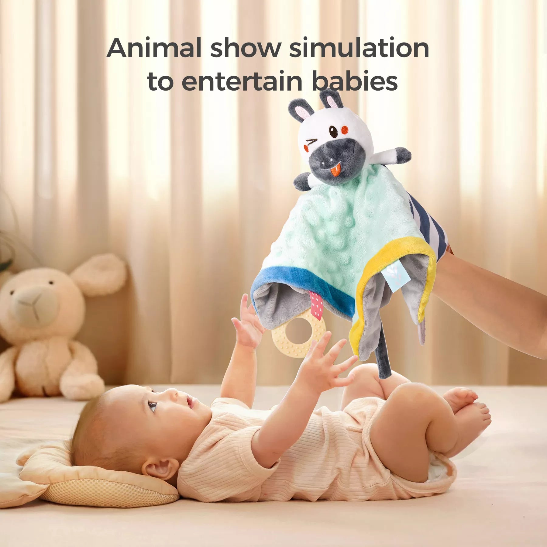 Baby lying on a pillow reaching up toward a plush animal security blanket lovey being held like a puppet. The blanket is soft with green, yellow, and blue fabric, and features a smiling animal head with attached teethers. Text reads: “Animal show simulation to entertain babies.”