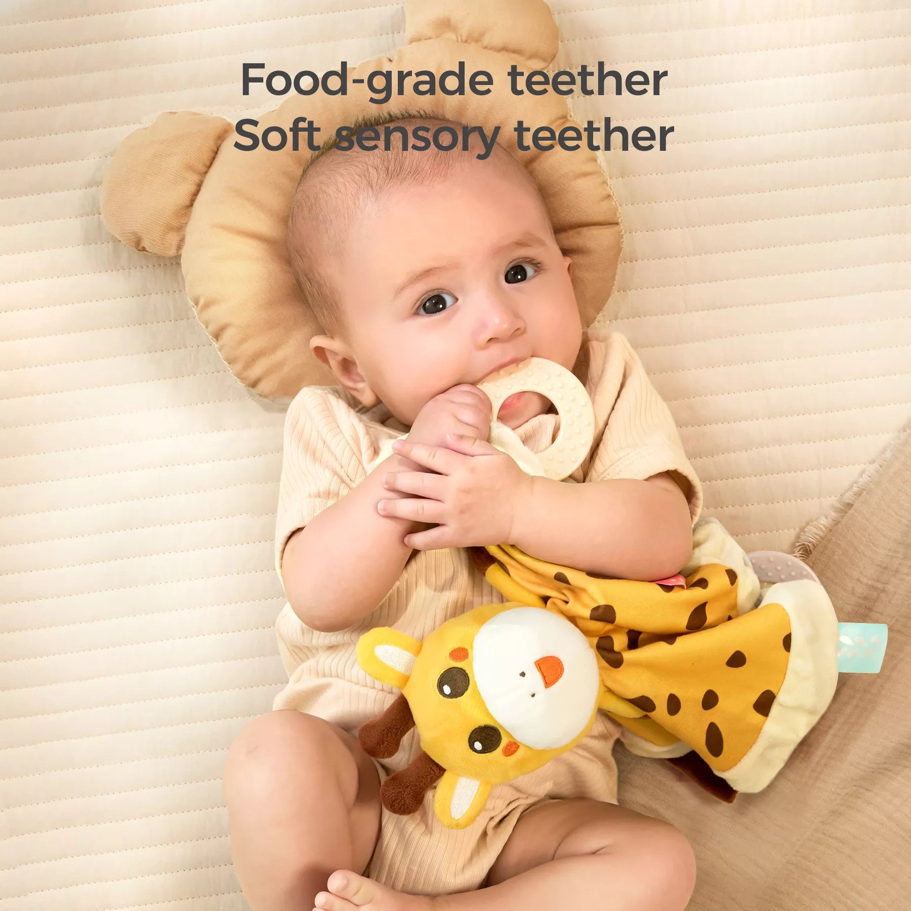Baby lying on a beige cushion, holding a giraffe-themed security blanket lovey. The baby is chewing on the attached beige food-grade teether ring, while the soft plush giraffe head and yellow spotted blanket body are visible. Text overlay reads “Food-grade teether, Soft sensory teether.”