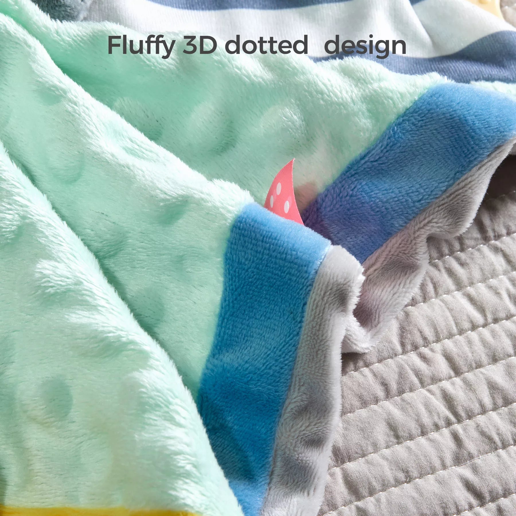 Close-up of a baby security blanket with a fluffy 3D dotted texture in soft mint green fabric, bordered with blue, yellow, and gray edges. A small pink polka-dot tag is visible for tactile play. Text above reads: “Fluffy 3D dotted design.”