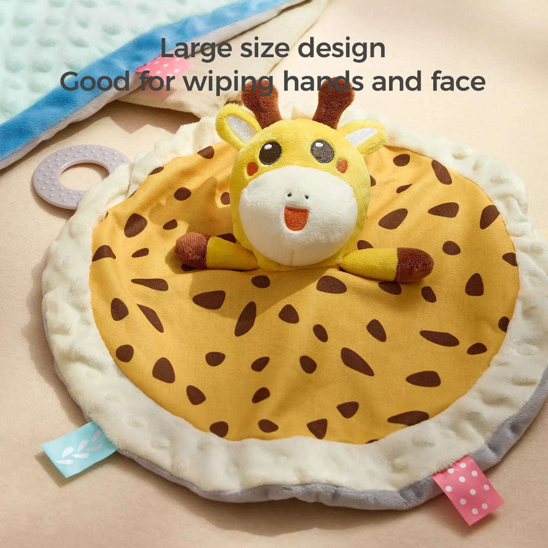 Round giraffe-themed security blanket lovey with a plush giraffe head in the center and a yellow spotted fabric body with brown accents. The blanket has a white fluffy edge, colorful fabric tags, and an attached teether. Text overlay reads “Large size design, Good for wiping hands and face.”