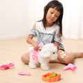 Young girl playing on the floor with a white walking and barking plush dog toy dressed in pink, using grooming accessories like a brush, stethoscope, hairdryer, and food bowl.
