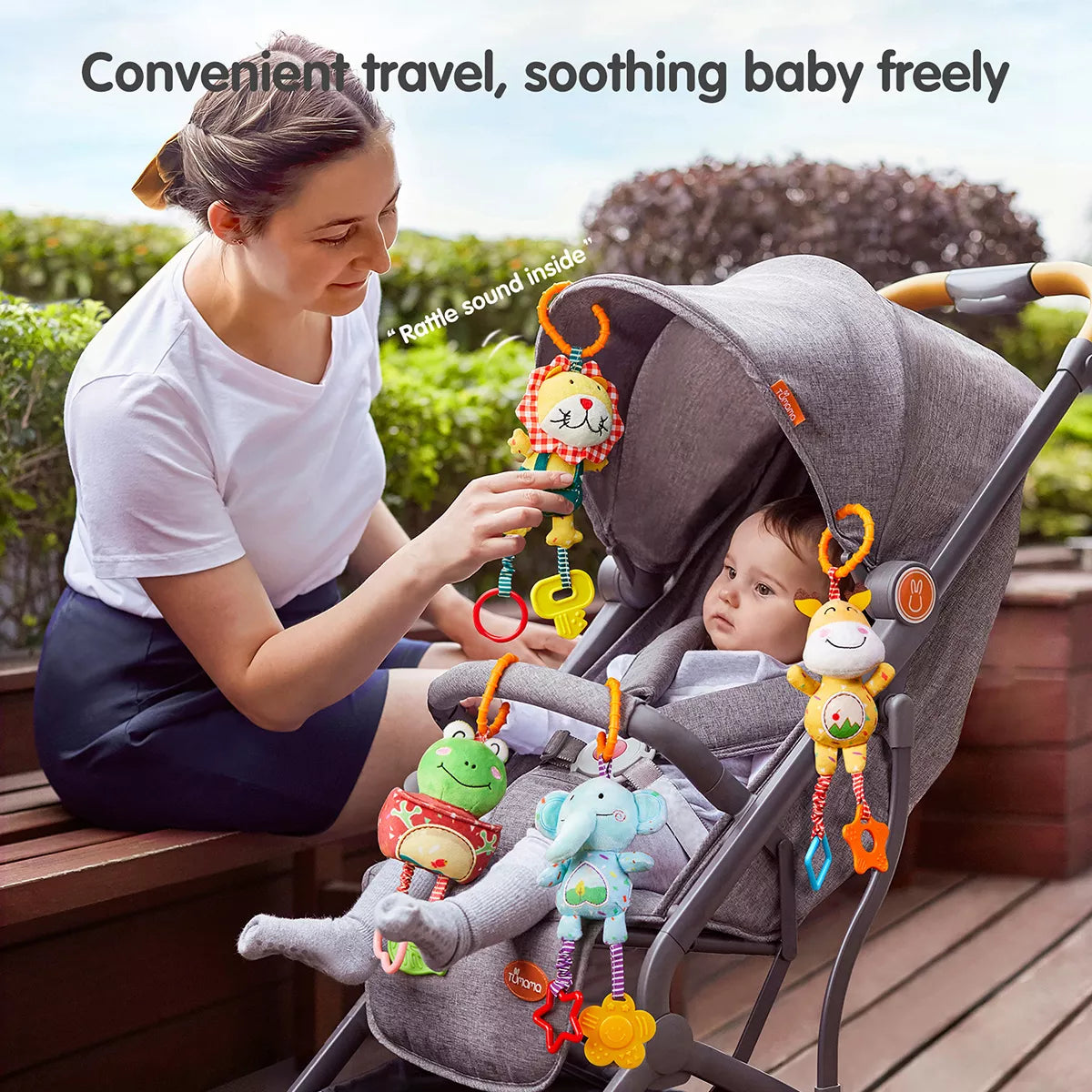 Mother adjusting a plush lion rattle hanging from a gray stroller canopy while a baby sits inside with three other animal rattles—a frog, elephant, and giraffe—clipped around the stroller’s seat, each toy featuring dangling rings and teethers
